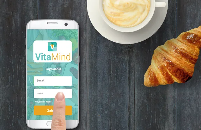 Smartfon z ekranem logowania aplikacji VitaMind obok kawy i rogalika