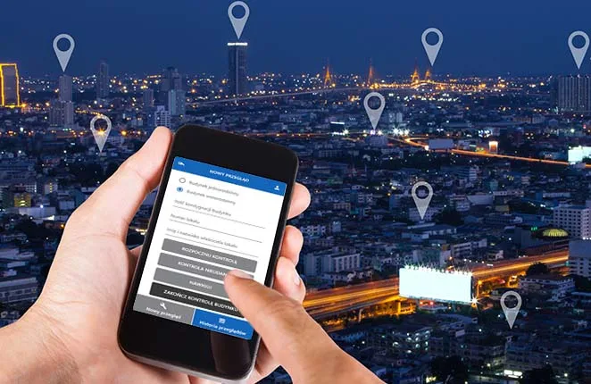 Smartfon z aplikacją Kominiarczyk w dłoni na tle miasta nocą z pinezkami lokalizacji